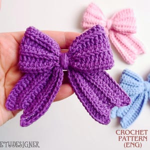 Op de afbeelding: Een close-up van een handgemaakte, paarse gehaakte strik die in een hand wordt gehouden, met andere gehaakte strikken in roze en blauw op de achtergrond. De tekst "CROCHET PATTERN (ENG)" is zichtbaar in de rechterbenedenhoek.