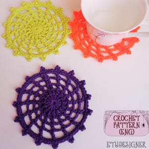 May include: Three colourful, handmade crochet coasters in yellow, orange, and purple. The coasters feature a circular design with an openwork pattern. One orange coaster supports a white mug. The image includes a logo that says "CROCHET PATTERN (ENG)" and "ETUDESIGNER."