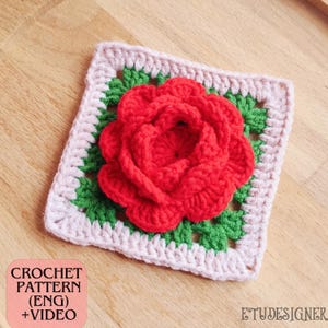 Puede incluir: Un cuadrado de ganchillo con una rosa roja en el centro, rodeada de ganchillo verde y blanco. El texto "CROCHET PATTERN (ENG) + VIDEO" es visible. Colocado sobre una superficie de madera.