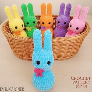 May include: A collection of colorful, handmade crochet bunny rabbits. The bunnies are in yellow, green, orange, purple, and pink, with black eyes and small bows. One blue bunny is in the foreground. The bunnies are in a woven basket. The text "CROCHET PATTERN (ENG)" is visible.