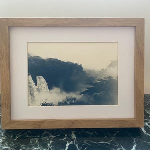 May include: A framed landscape print depicting a misty mountain scene in blue and white tones. The artwork is presented with a white mount and a light brown wooden frame. The frame is displayed on a dark green marble surface, offering a classic look.
