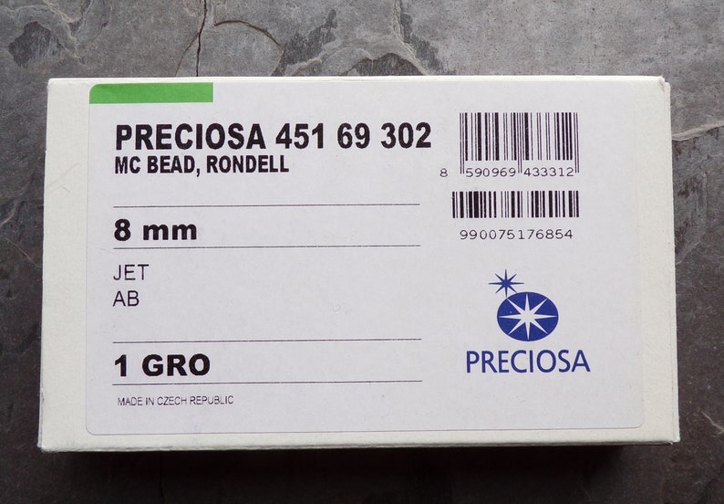 Puede incluir: Una caja blanca con el texto "PRECIOSA 451 69 302 MC BEAD, RONDELL" y "8 mm". La caja tambi&eacute;n incluye el texto "JET AB" y "1 GRO". El logotipo de Preciosa es visible. Fabricado en la Rep&uacute;blica Checa.