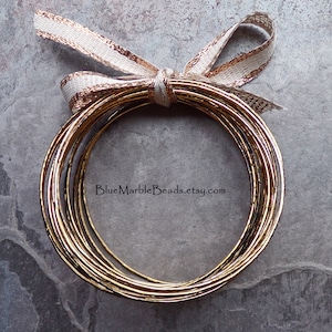 May include: A stack of gold-colored bangles tied with a cream and gold ribbon. The bangles have a textured surface. The Etsy shop name "BlueMarbleBeads.etsy.com" is visible in the image.