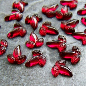 May include: A selection of small, ruby-red glass leaf-shaped beads. Each bead features a detailed leaf vein design and a glossy finish. The beads are scattered on a grey, textured surface, creating a visually appealing arrangement.