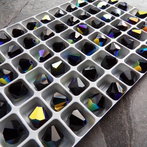 Puede incluir: Una bandeja blanca llena de cuentas de cristal negro facetadas. Cada cuenta tiene m&uacute;ltiples facetas que reflejan un arco&iacute;ris de colores. Las cuentas est&aacute;n dispuestas en un patr&oacute;n de cuadr&iacute;cula, creando una exhibici&oacute;n visualmente atractiva.