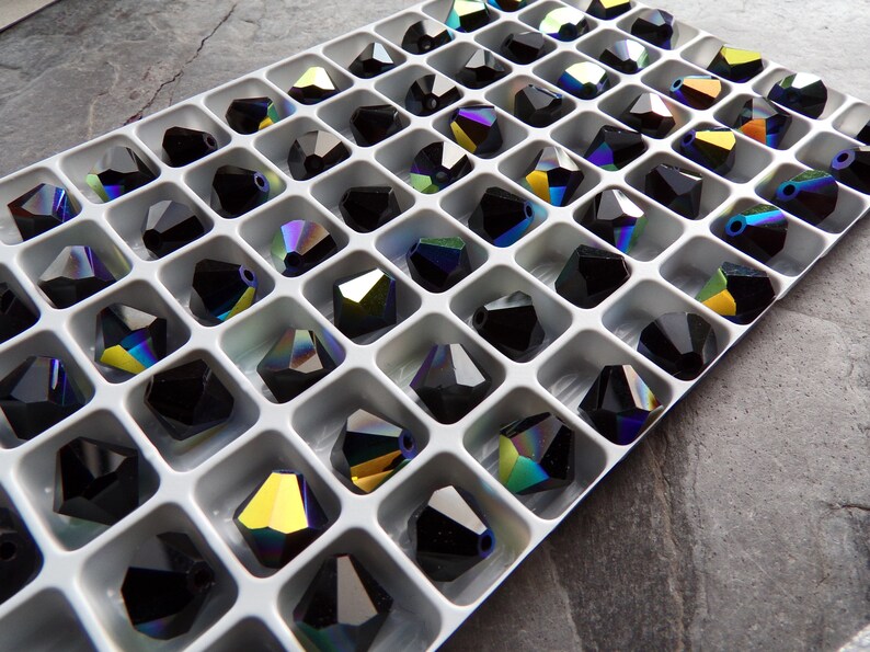 Puede incluir: Una bandeja blanca llena de cuentas de cristal negro facetadas. Cada cuenta tiene m&uacute;ltiples facetas que reflejan un arco&iacute;ris de colores. Las cuentas est&aacute;n dispuestas en un patr&oacute;n de cuadr&iacute;cula, creando una exhibici&oacute;n visualmente atractiva.