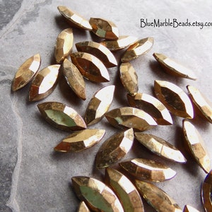 May include: A collection of gold-toned, faceted, marquise-shaped glass beads. The beads are scattered on a gray, textured surface.