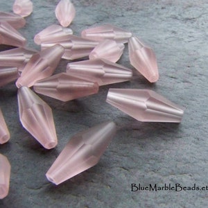 May include: A selection of pale pink, elongated beads with faceted sides. The beads have a frosted finish and are spread across a grey surface. These beads are likely for jewellery making or other craft projects.