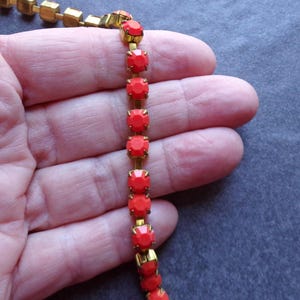 Puede incluir: Una pulsera dorada con gemas rojas vibrantes. Las gemas son de corte cuadrado y están engastadas en un marco de metal dorado. La pulsera se muestra sobre un fondo neutro.