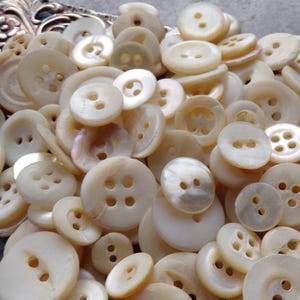 May include: A collection of off-white buttons in various sizes and shapes. The buttons have two or four holes, and some have a pearlescent sheen. The buttons are scattered, creating a textured and detailed display.
