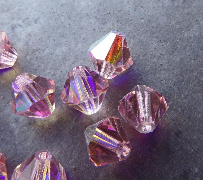 May include: A group of iridescent pink faceted glass beads. The beads have a rainbow sheen and are shaped like a pyramid.