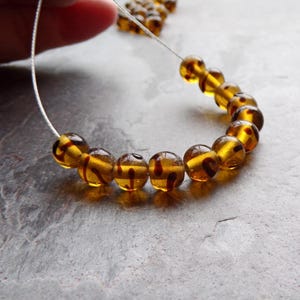 May include: A string of amber-coloured, round beads with dark brown veining. The beads are strung on a thin, silver-coloured wire. The beads are approximately 1.3 cm in diameter. The background is a grey, textured surface.