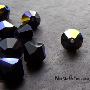 Puede incluir: Cuentas negras facetadas con reflejos iridiscentes. Las cuentas tienen forma geom&eacute;trica y un agujero para ensartar. Las cuentas est&aacute;n dispersas sobre una superficie gris. La direcci&oacute;n del sitio web "BlueMarbleBeads.etsy.com" es visible.