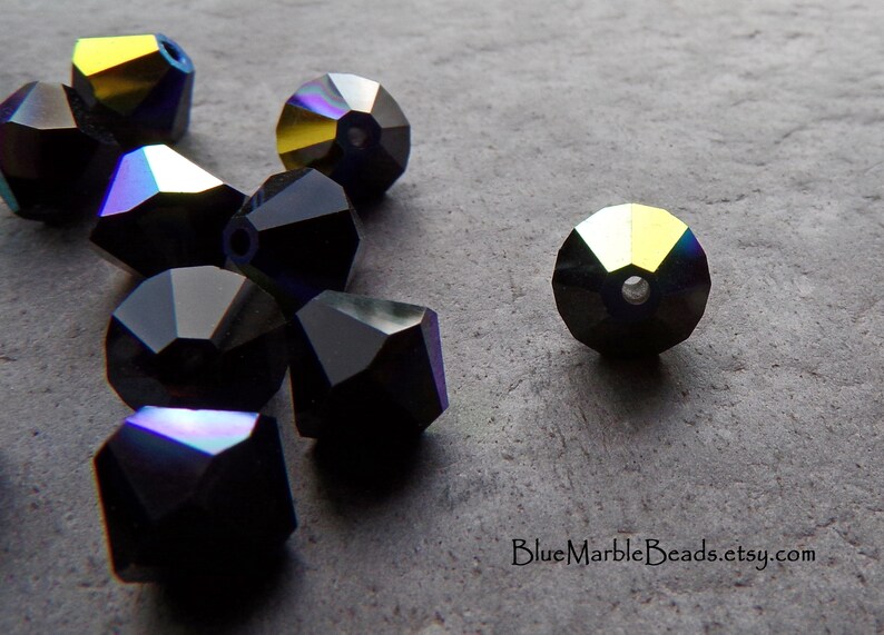 Puede incluir: Cuentas negras facetadas con reflejos iridiscentes. Las cuentas tienen forma geom&eacute;trica y un agujero para ensartar. Las cuentas est&aacute;n dispersas sobre una superficie gris. La direcci&oacute;n del sitio web "BlueMarbleBeads.etsy.com" es visible.