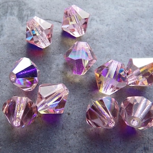 May include: A group of ten pink iridescent faceted glass beads. The beads are shaped like a pyramid with a square base. The beads are all the same size and have a shiny, reflective surface.