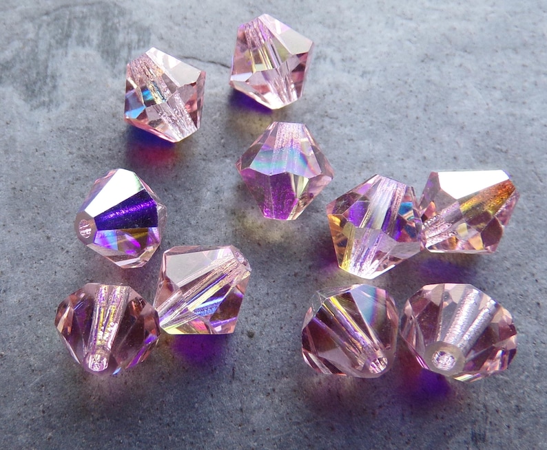 May include: A group of ten pink iridescent faceted glass beads. The beads are shaped like a pyramid with a square base. The beads are all the same size and have a shiny, reflective surface.