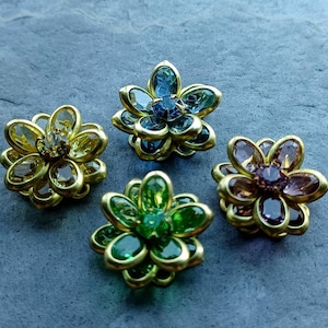 May include: Four gold-toned flower-shaped brooches with jeweled centers. The brooches feature different colored jewels: blue, green, yellow, and purple.