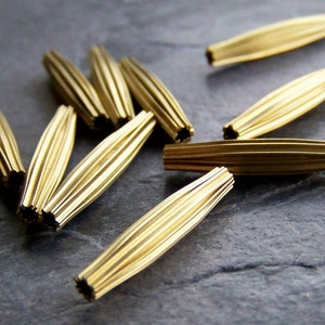 Könnte beinhalten: Nahaufnahme von mehreren goldfarbenen, geriffelten Metallperlen. Die Perlen sind länglich mit einer Reihe paralleler Linien entlang ihrer Länge. Die Enden der Perlen haben ein sternförmiges Muster. Die Perlen sind auf einer dunkelgrauen Oberfläche verstreut.