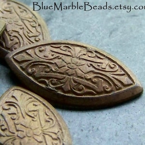 May include: Close-up of several antique bronze-colored oval beads. Each bead features a detailed floral and scrollwork design. The beads are arranged on a textured surface. The Etsy shop name "BlueMarbleBeads.etsy.com" is visible in the image.