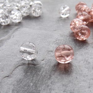 May include: Close-up of clear and rose-colored glass beads. The clear beads are clustered together, while the rose-colored beads are grouped separately. Two individual beads, one clear and one rose, are in the foreground. The beads have a faceted, textured appearance.