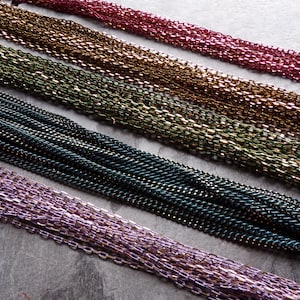 May include: An assortment of colorful chains in shades of red, gold, green, teal, and purple. The chains are arranged in parallel lines, creating a textured and visually appealing display. The chains appear to be made of small, interconnected links.