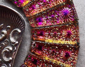May include: A collection of seven rectangular glass beads arranged in a fan shape, each featuring a vibrant iridescent finish with a repeating starburst pattern in shades of magenta, gold, and purple, alongside a metallic decorative tray.