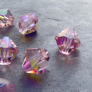 May include: A group of pink iridescent glass beads with a faceted design. The beads are scattered on a grey surface.