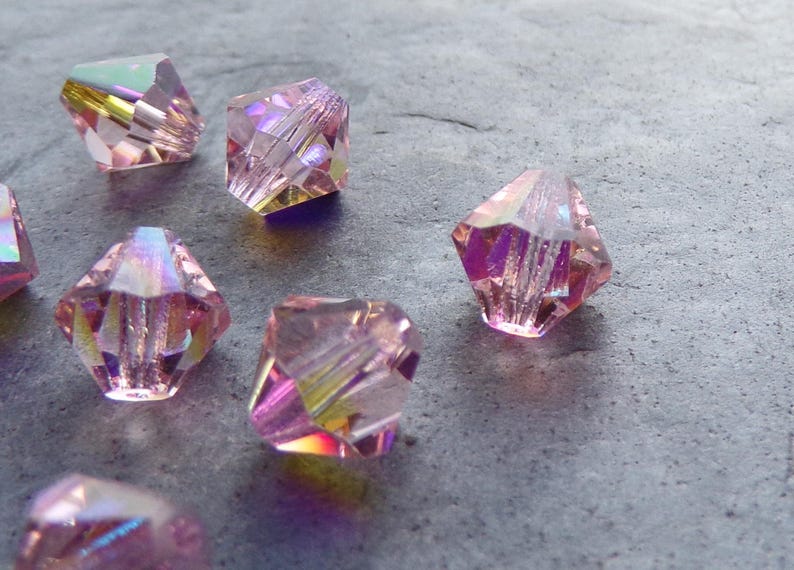 May include: A group of pink iridescent glass beads with a faceted design. The beads are scattered on a grey surface.