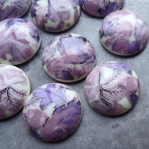 May include: Assortment of round, purple and white cabochons with a floral design. The cabochons have a glossy finish and are decorated with silver glitter.