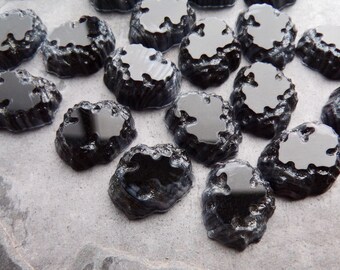 May include: A collection of small, irregularly shaped, black glass pieces. Each piece has a textured, rough exterior and a smooth, reflective surface. The pieces are scattered across a gray, textured surface, creating a visually interesting pattern.