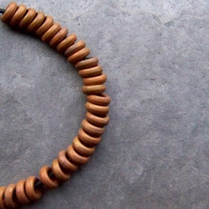 May include: A curved section of a necklace featuring numerous small, round, wooden beads. The beads are a warm, light brown color and strung on a dark cord. The background is a textured, gray surface.