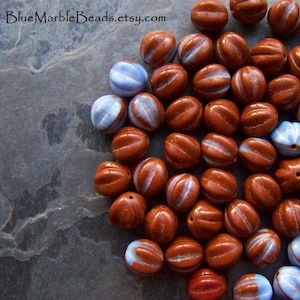 Puede incluir: Una colección de cuentas de vidrio marrón y blanco con forma de grano de café. Las cuentas tienen una franja blanca que recorre el centro de cada cuenta.