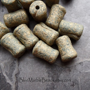May include: A pile of green and gold ceramic beads with a rough, textured surface. The beads are cylindrical in shape and have a metallic sheen.