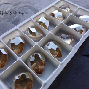 May include: A close-up of a white plastic container filled with 12 clear glass teardrop-shaped beads with a gold iridescent finish. The beads are arranged in rows and are all the same size.