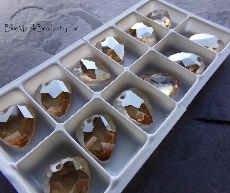 May include: A close-up of a white plastic container filled with 12 clear glass teardrop-shaped beads with a gold iridescent finish. The beads are arranged in rows and are all the same size.