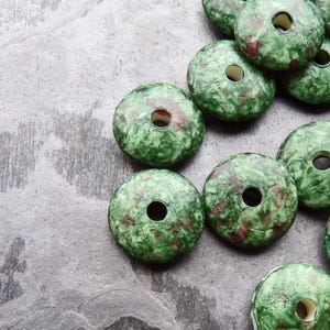 May include: A collection of green and maroon ceramic beads with a central hole. The beads have a mottled, textured appearance, and are arranged on a gray, textured surface. These beads are suitable for jewelry making and crafts.