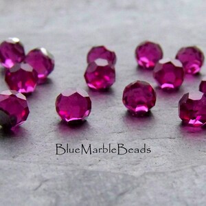 May include: A close-up of a group of 15 small, faceted, round, magenta-coloured glass beads. The beads are scattered on a grey, textured surface. The text "BlueMarbleBeads" is visible in the lower right corner of the image.