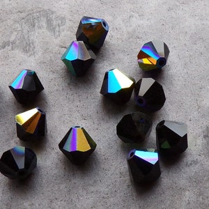 Puede incluir: Un conjunto de cuentas negras facetadas con superficies iridiscentes de colores arco&iacute;ris. Las cuentas tienen forma geom&eacute;trica y est&aacute;n dispersas sobre una superficie gris. Adecuadas para la creaci&oacute;n de joyas.