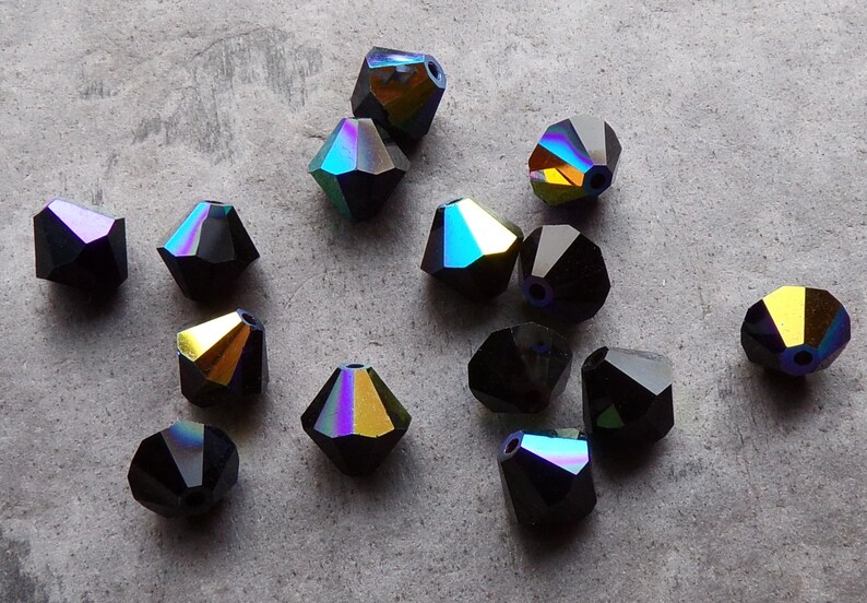 Puede incluir: Un conjunto de cuentas negras facetadas con superficies iridiscentes de colores arco&iacute;ris. Las cuentas tienen forma geom&eacute;trica y est&aacute;n dispersas sobre una superficie gris. Adecuadas para la creaci&oacute;n de joyas.