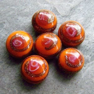 May include: Six orange, round beads with a spiral design. Each bead has a red and white swirl in the center, surrounded by a glittery brown band. The beads are arranged on a gray surface.