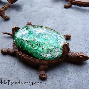 May include: A brown turtle-shaped figurine featuring a large, oval-shaped cabochon with a vibrant green and silver metallic, crackled texture set into its shell. The turtle has a textured, scaly surface and is positioned on a gray surface.