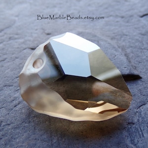 May include: A clear, faceted, teardrop-shaped crystal bead with a hole drilled through the top. The bead has a light brown color and is lying on a gray surface.
