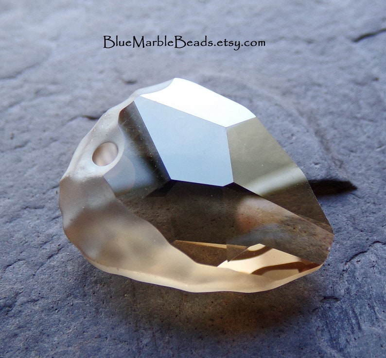 May include: A clear, faceted, teardrop-shaped crystal bead with a hole drilled through the top. The bead has a light brown color and is lying on a gray surface.