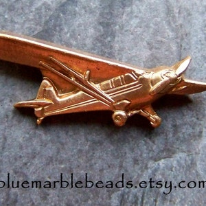 May include: A small, detailed, copper-colored airplane pin. The pin features a propeller, wings, and a fuselage. The Etsy shop URL bluemarblebeads.etsy.com is visible at the bottom of the image.