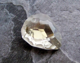 May include: A faceted, teardrop-shaped crystal pendant with a pale yellow hue. The crystal has multiple facets that reflect light, creating a sparkling effect. The pendant is set against a gray, textured stone surface.