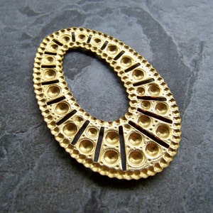 May include: A gold-colored, oval-shaped metal embellishment. The piece features a detailed design with small circles and vertical cutouts along the edges. The center is open, and the overall aesthetic is ornate and decorative.