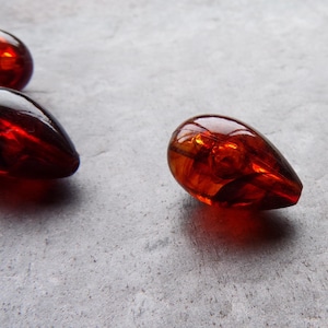 May include: Several teardrop-shaped, amber-colored beads are scattered on a gray surface. The beads have a glossy, reflective surface, with light refracting through them. The overall effect is one of warmth and depth, suggesting a collection of decorative elements.