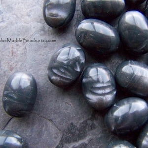 May include: A close-up of a group of oval-shaped, dark gray, iridescent glass beads. The beads have a smooth, polished surface and a subtle shimmer.