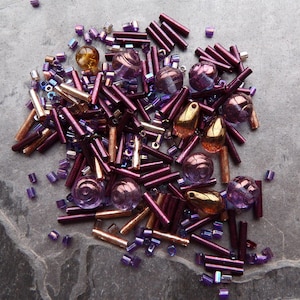 Puede incluir: Una colección de cuentas de varios tipos y colores: morado, dorado y bronce. El surtido incluye cuentas cilíndricas, redondas y en forma de lágrima, además de pequeñas cuentas cuadradas. Las cuentas están sobre una superficie gris texturizada.