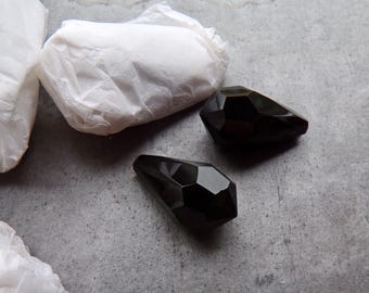 May include: Three faceted black gemstone beads, teardrop-shaped, are shown on a grey surface. White tissue paper is partially visible in the background. The beads have a polished, reflective surface.